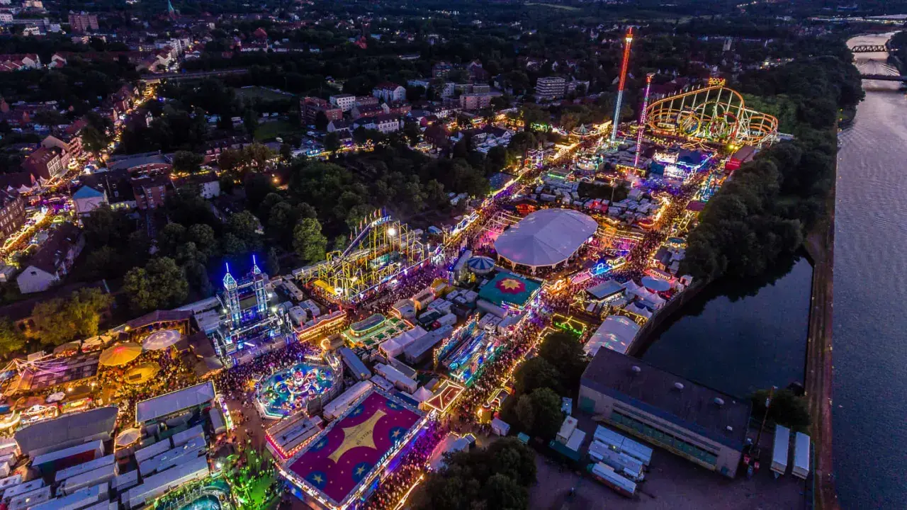 Luftaufnahme eines beleuchteten Volksfestes mit Fahrgeschäften, Zelten und Menschenmassen. Ein typisches Bild für Veranstaltungen NRW.