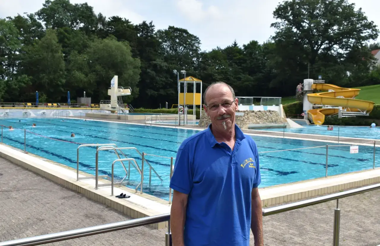 Ein Mann steht vor dem Freibad Bad Driburg. Im Hintergrund sind Schwimmer, Rutschen und ein Sprungturm zu sehen.
