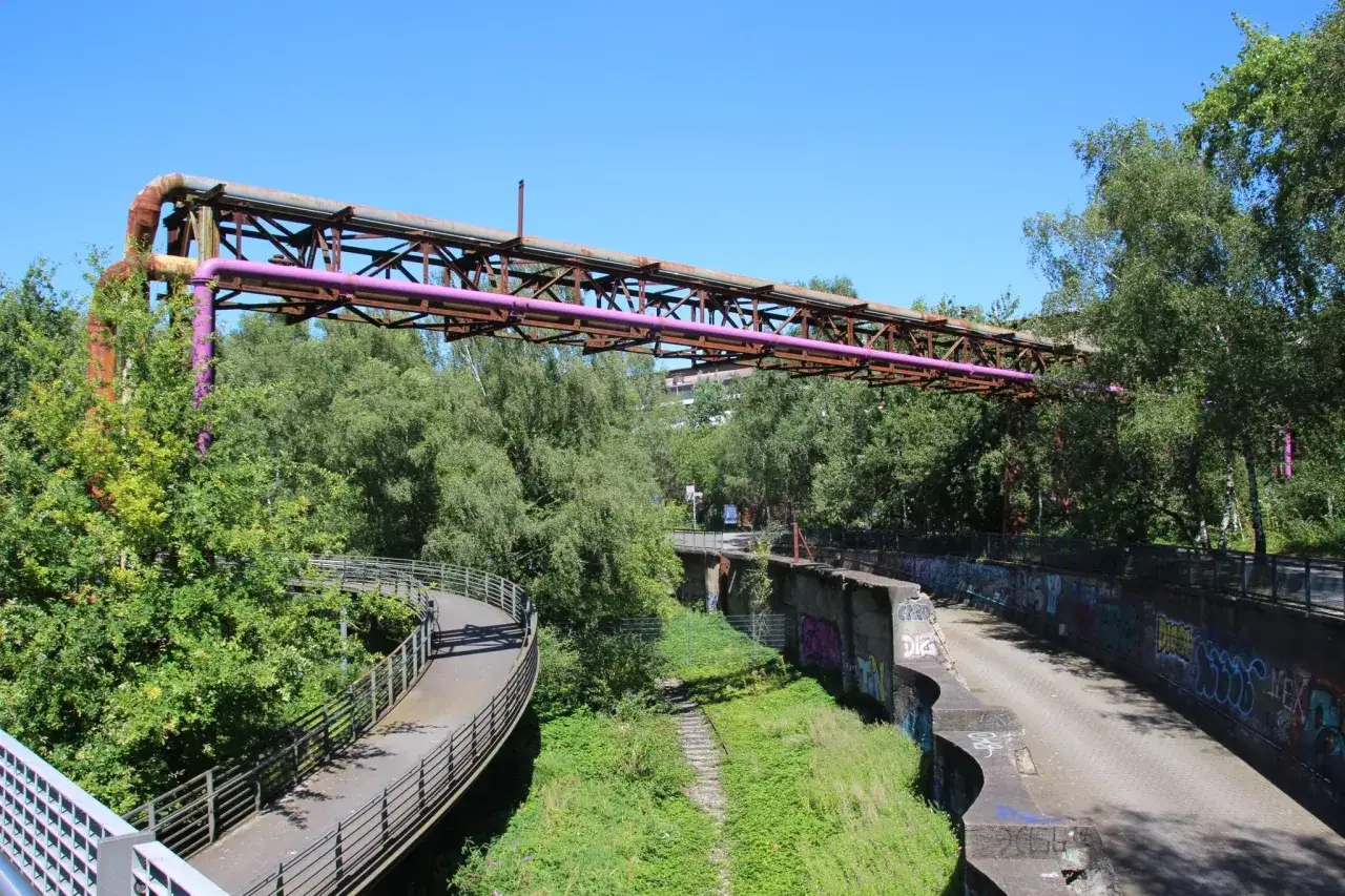 Alte Rohrleitungen der Erzbahntrasse Bochum ragen &uuml;ber &uuml;ppiges Gr&uuml;n. Ein geschwungener Weg f&uuml;hrt durch die Natur.