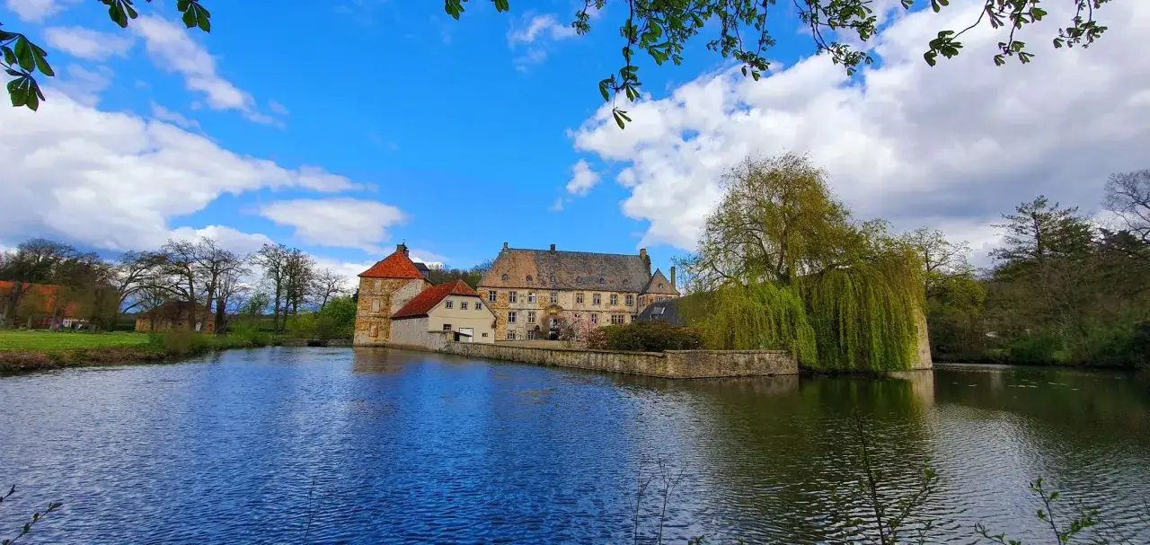 Wasserschloss in Halle Westfalen, umgeben von einem Teich und grünen Bäumen, unter blauem Himmel mit Wolken.