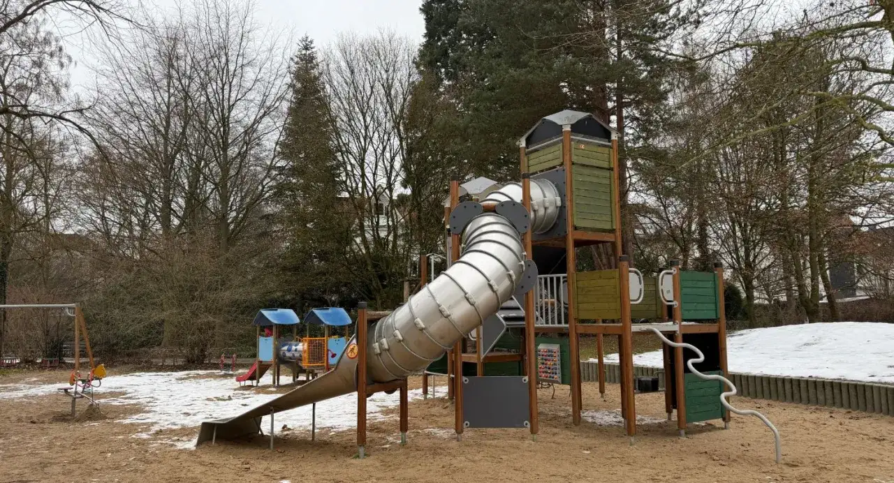 Ein Spielplatz im Herford Park mit einer gro&szlig;en R&ouml;hrenrutsche und Schaukeln, umgeben von kahlen B&auml;umen und etwas Schnee.