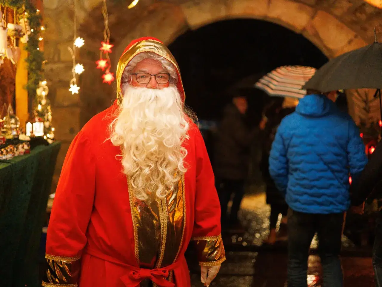Weihnachtsmann auf einem Markt in Tecklenburg. Lichterketten und ein Stand mit Weihnachtsdeko im Hintergrund.