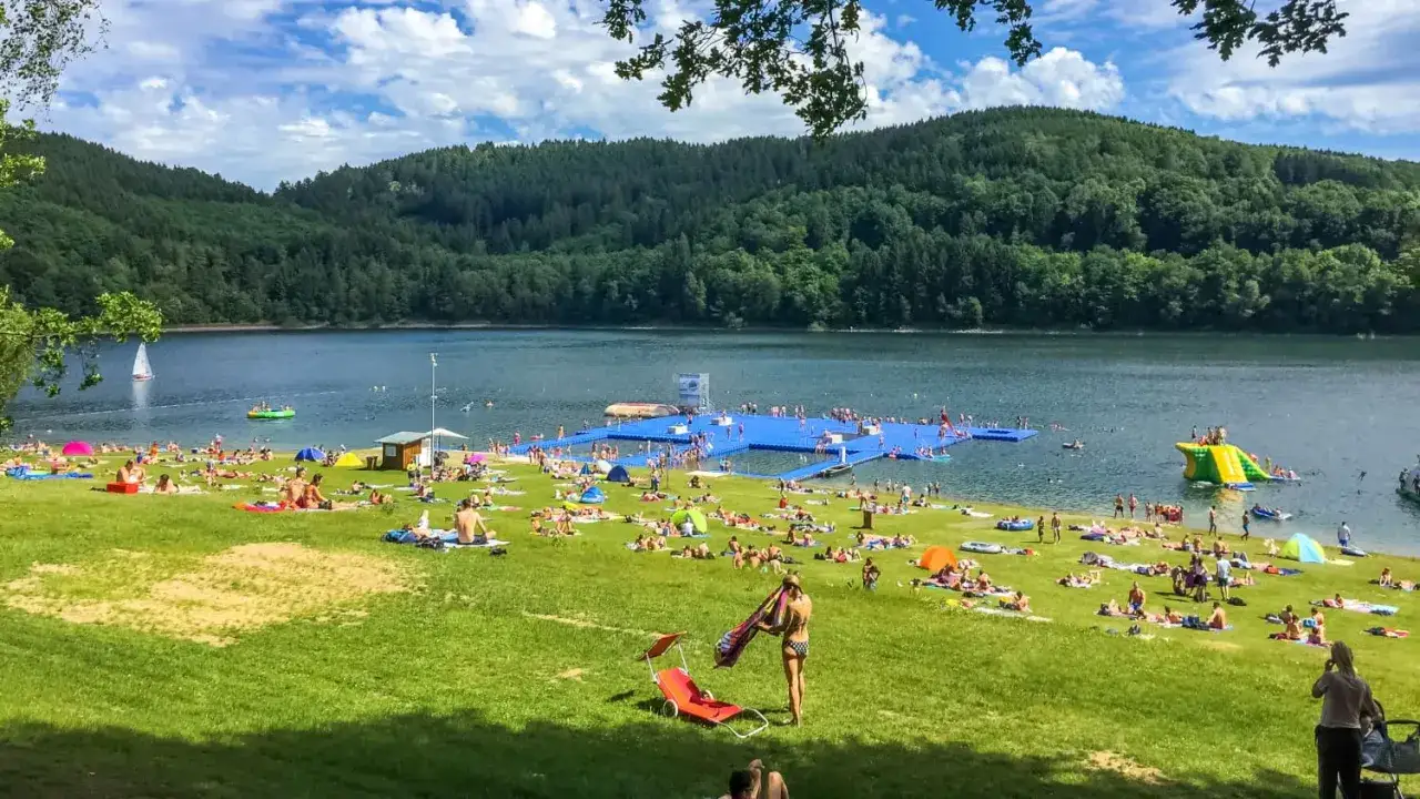 Sonniger Tag am Strand NRW. Viele Menschen entspannen sich auf der gr&uuml;nen Wiese, baden im See oder genie&szlig;en die schwimmende Plattform mit Rutsche.