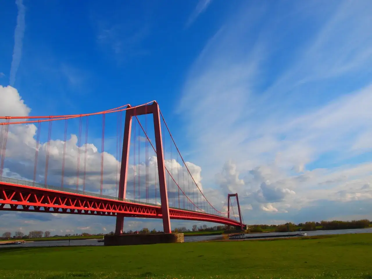 Die rote Rheinbr&uuml;cke bei Emmerich ragt unter einem blauen Himmel mit Wolken auf. Die Rheinpromenade l&auml;dt zum Spaziergang ein.