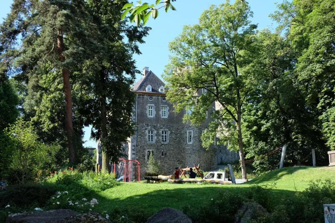 Ein Steinhaus mit T&uuml;rmchen und Fensterl&auml;den, umgeben von gr&uuml;nen B&auml;umen. Im Vordergrund ein Spielplatz mit Holzst&auml;mmen und einem Fu&szlig;balltor im Frankenberger Park.