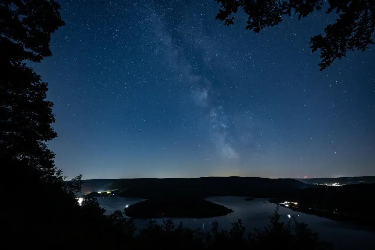Ein atemberaubender Sternenhimmel über einem See in NRW. Die Milchstraße funkelt, während Lichter am Ufer glitzern. Perfekt zum Sterne gucken in NRW.