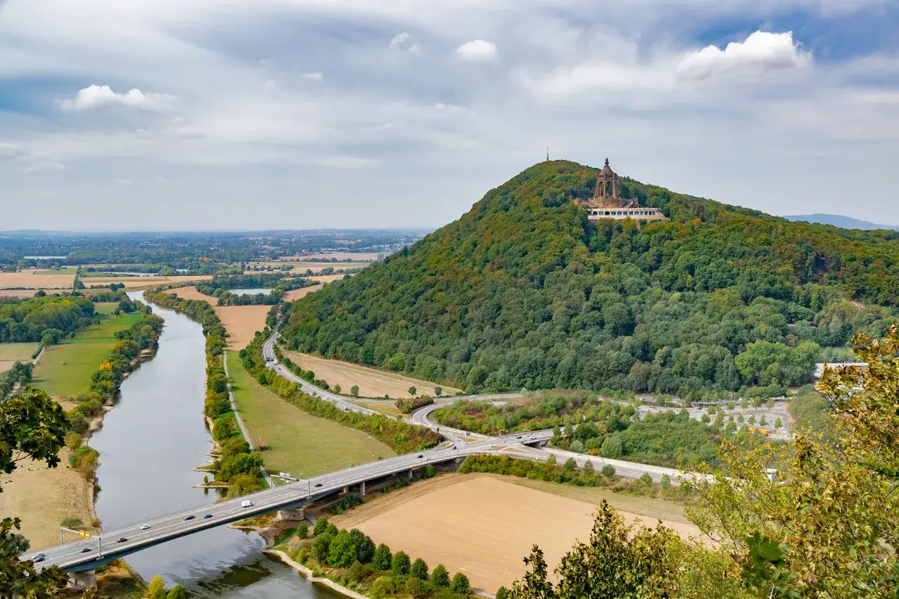 Blick auf Porta Westfalica mit dem Kaiser-Wilhelm-Denkmal auf dem Berg. Ein Fluss schlängelt sich durch die Landschaft, über den eine Brücke führt.