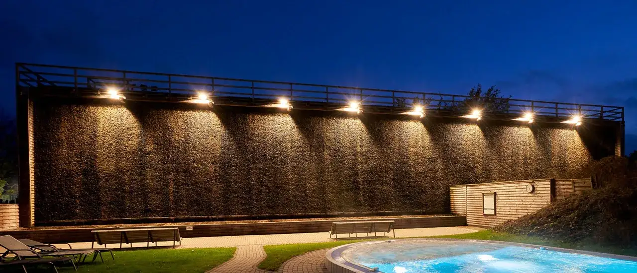 Abendliche Szene an der Therme Sauerland: Ein gro&szlig;es Gradierwerk mit beleuchteten Reisigw&auml;nden, davor ein Pool und Sitzb&auml;nke.
