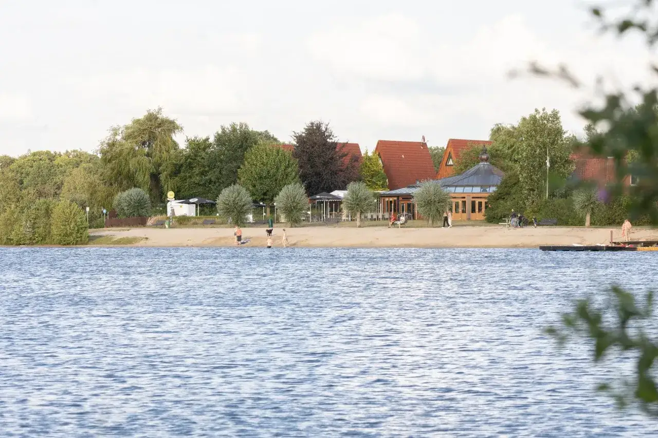 Ein sonniger Tag am See in Gronau. Menschen genießen den Strand und das Wasser, während im Hintergrund Gebäude und Bäume zu sehen sind.