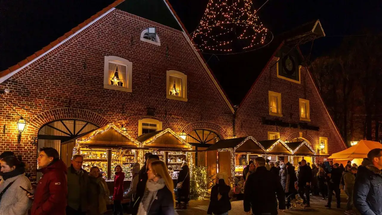 Besucher bummeln &uuml;ber den velen Waldweihnachtsmarkt, vorbei an festlich beleuchteten Buden und Backsteinh&auml;usern. Ein Lichterbaum funkelt.
