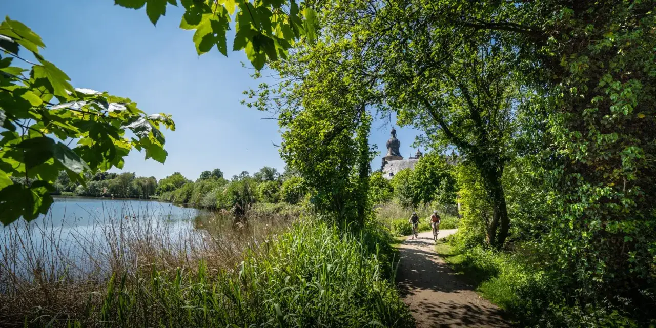Zwei Radfahrer genießen die hohe Mark Radroute entlang eines Sees, im Hintergrund ein Schloss.