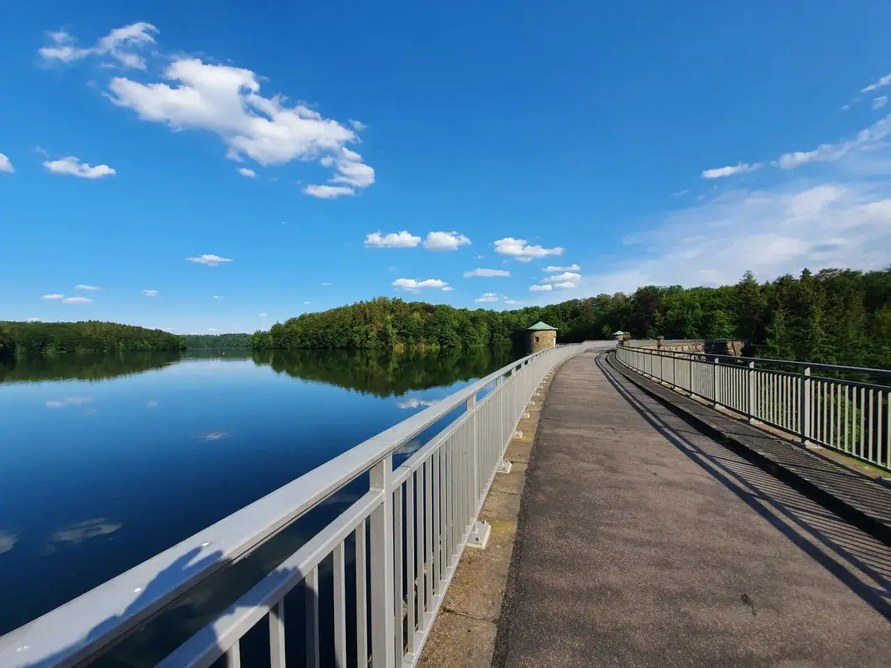 Der Weg entlang der Neyetalsperre lädt zu einem Spaziergang ein. Der blaue Himmel mit weißen Wolken spiegelt sich im ruhigen Wasser.