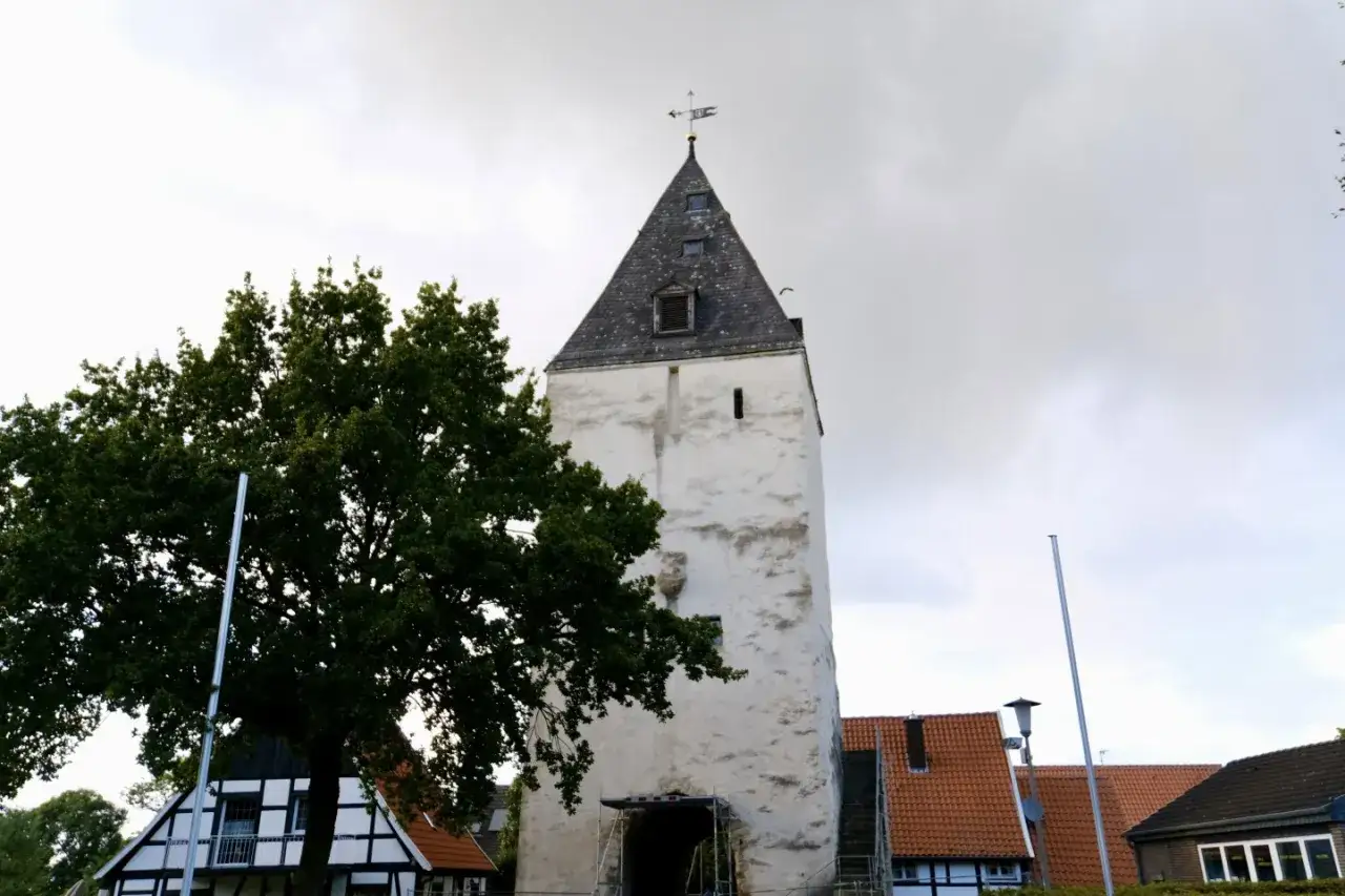 Der Stromberg in Oelde, ein alter Turm, ragt neben einem Baum und Fachwerkh&auml;usern in den Himmel.