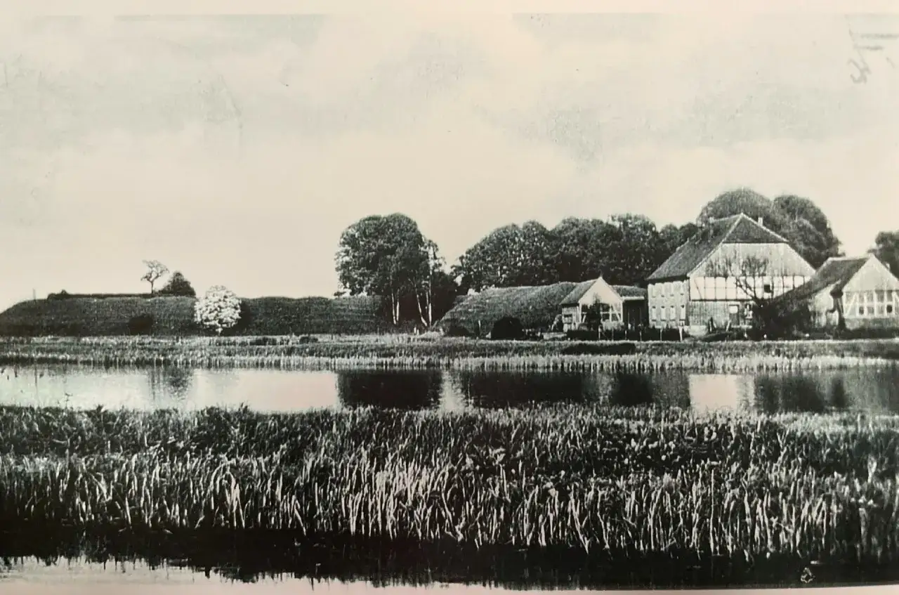 Idyllisches Bauernhaus mit Teich und Schilf, ein gut rietberg-Motiv, umgeben von B&auml;umen und einer sanften H&uuml;gellandschaft.