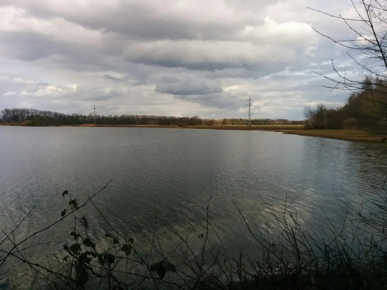 Ruhiger See mit B&auml;umen am Ufer. Ein Strommast ragt in den bew&ouml;lkten Himmel. Der boker kanal radweg l&auml;dt zum Erkunden ein.