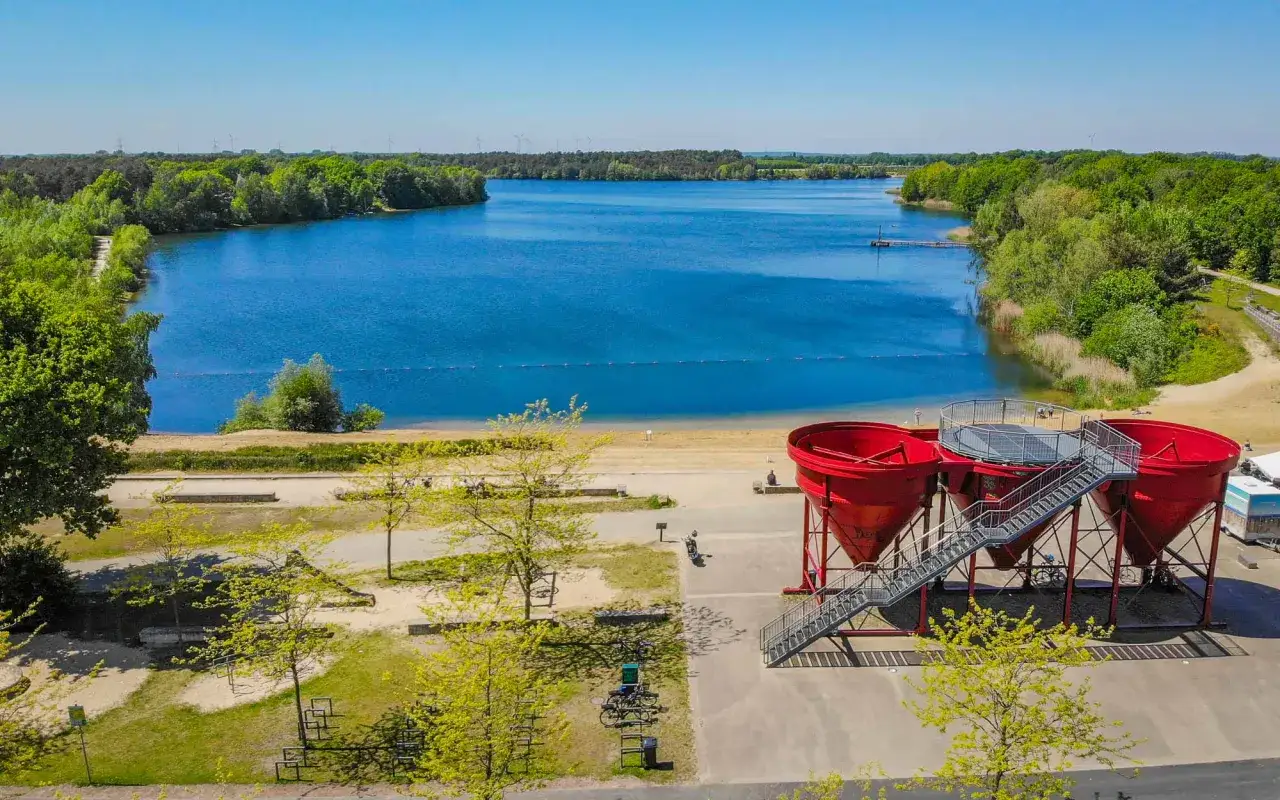 Der Offlumer See mit seinem tiefblauen Wasser, umgeben von gr&uuml;nen B&auml;umen. Im Vordergrund stehen drei rote Trichter, die wie riesige Becher aussehen.
