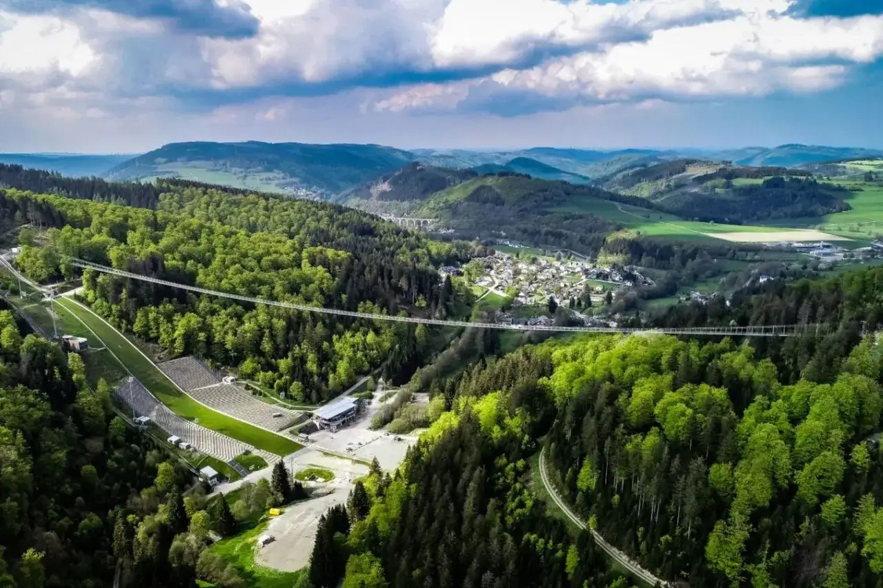 Der Willingen Skywalk überspannt ein grünes Tal mit Wäldern und einer kleinen Stadt. Eine Skisprungschanze ist links zu sehen.
