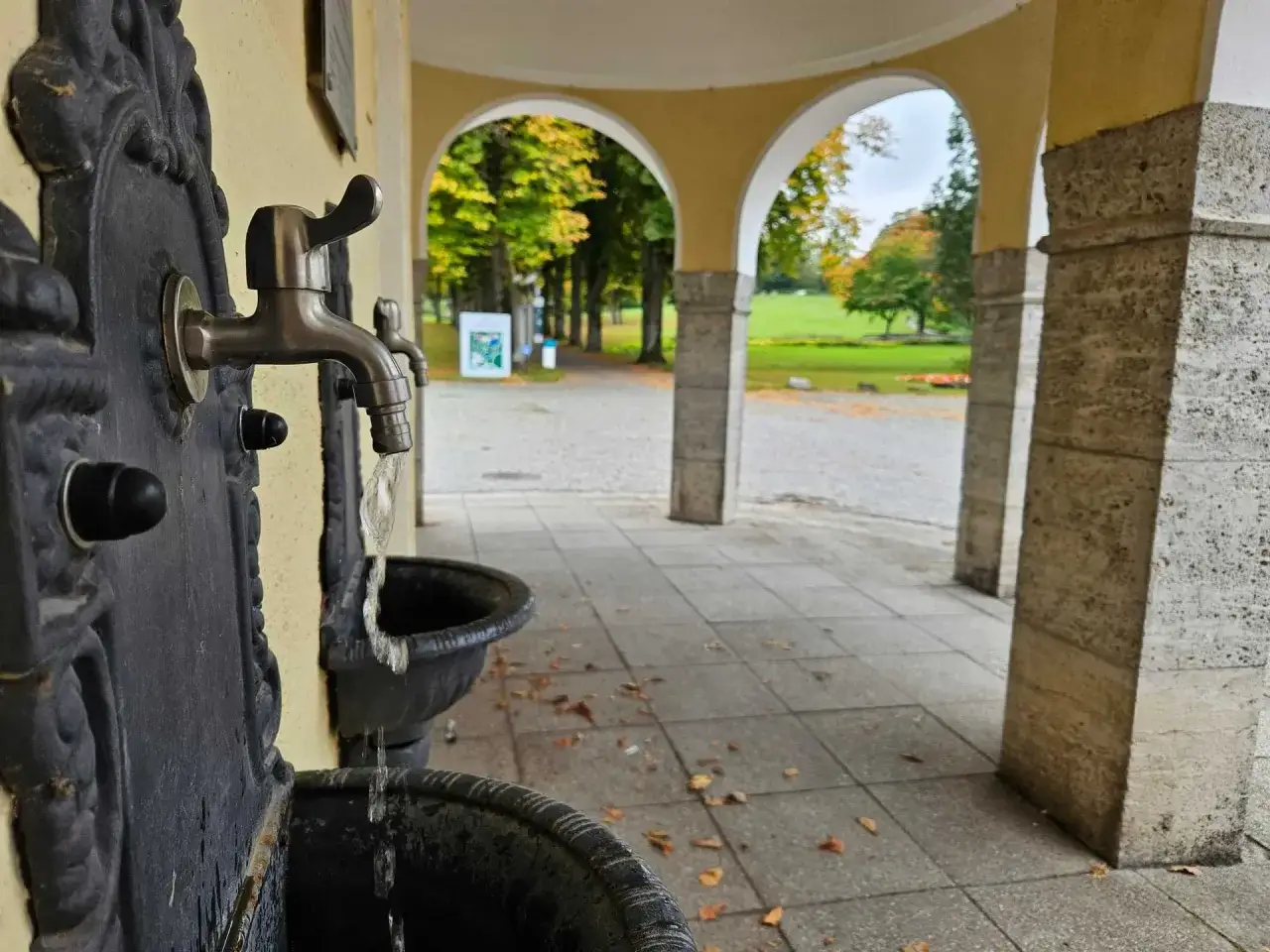 Erfrischendes Wasser fließt aus einem historischen Brunnen im Kurpark Bad Hermannsborn. Herbstlaub liegt auf dem gepflasterten Weg.