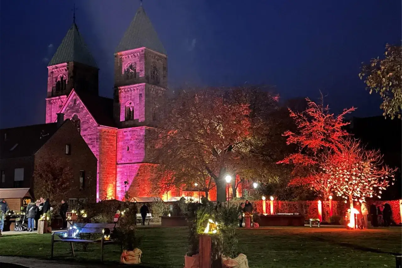 Festlicher weihnachtsmarkt legden vor einer rosa beleuchteten Kirche. B&auml;ume sind rot illuminiert, Menschen schlendern durch die festliche Atmosph&auml;re.