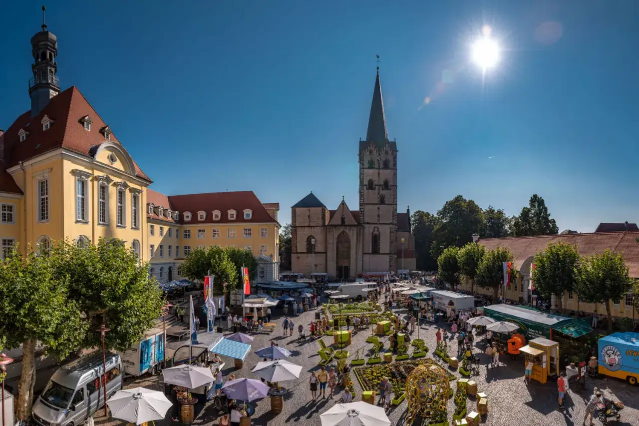 Bunte Marktst&auml;nde beleben die Herford Innenstadt. Menschen schlendern vorbei an der Kirche und dem gelben Rathaus unter strahlend blauem Himmel.
