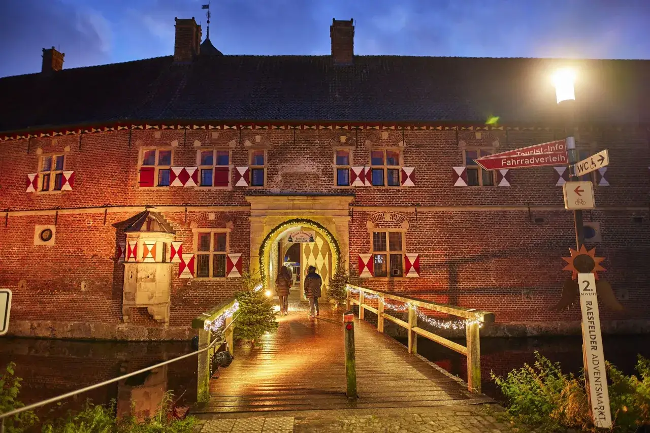 Ein beleuchteter Holzsteg f&uuml;hrt zum Wasserschloss, wo der Raesfelder Adventsmarkt mit Lichtern und Weihnachtsb&auml;umen lockt.