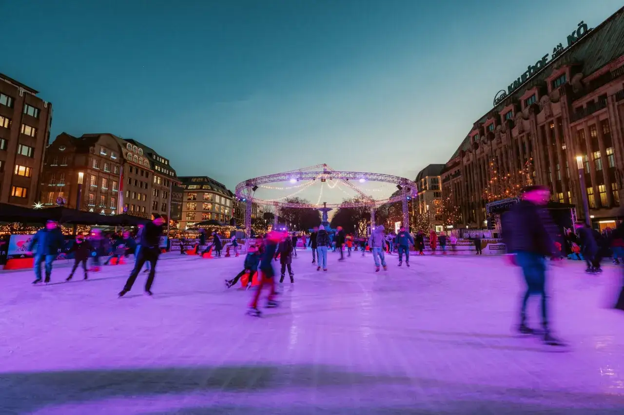 Ein belebter weihnachtsmarkt mit schlittschuhbahn, Menschen gleiten im Abendlicht über das Eis, umgeben von festlicher Beleuchtung und historischen Gebäuden.