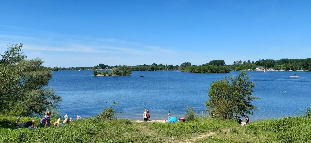 Sonniger Tag am Nesthauser See. Menschen entspannen am Ufer, Segelboote gleiten &uuml;ber das Wasser.