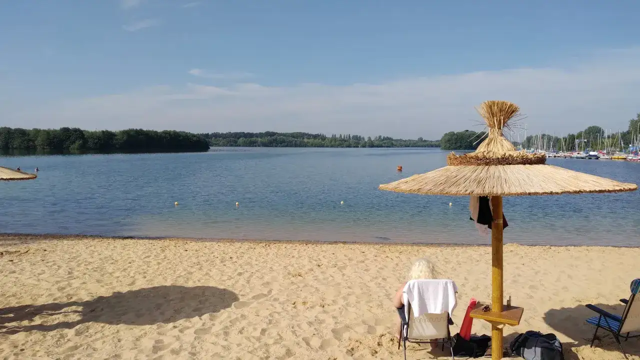 Ein sonniger Tag am Lippesee Paderborn. Eine Frau entspannt sich im Schatten eines Strohschirms am Sandstrand.