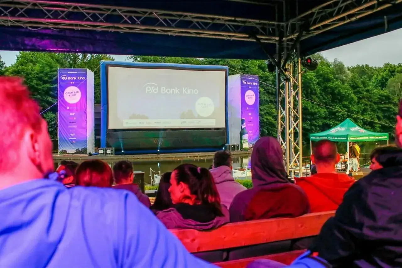 Menschen sitzen auf Holzbänken und genießen einen Film im freiluft kino. Eine große Leinwand mit dem Logo