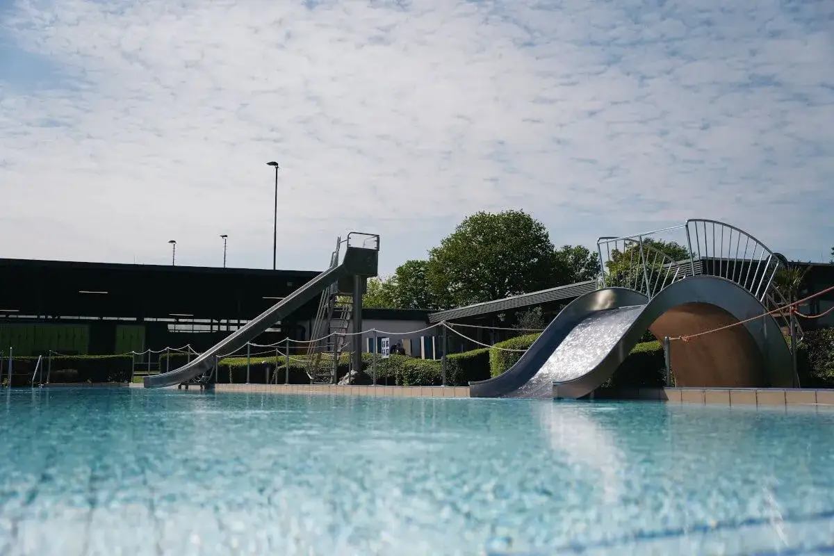 Wasserspaß im Freibad Elverdissen: Zwei Rutschen führen ins kühle Nass, umgeben von grünen Hecken und einem blauen Himmel mit Wolken.