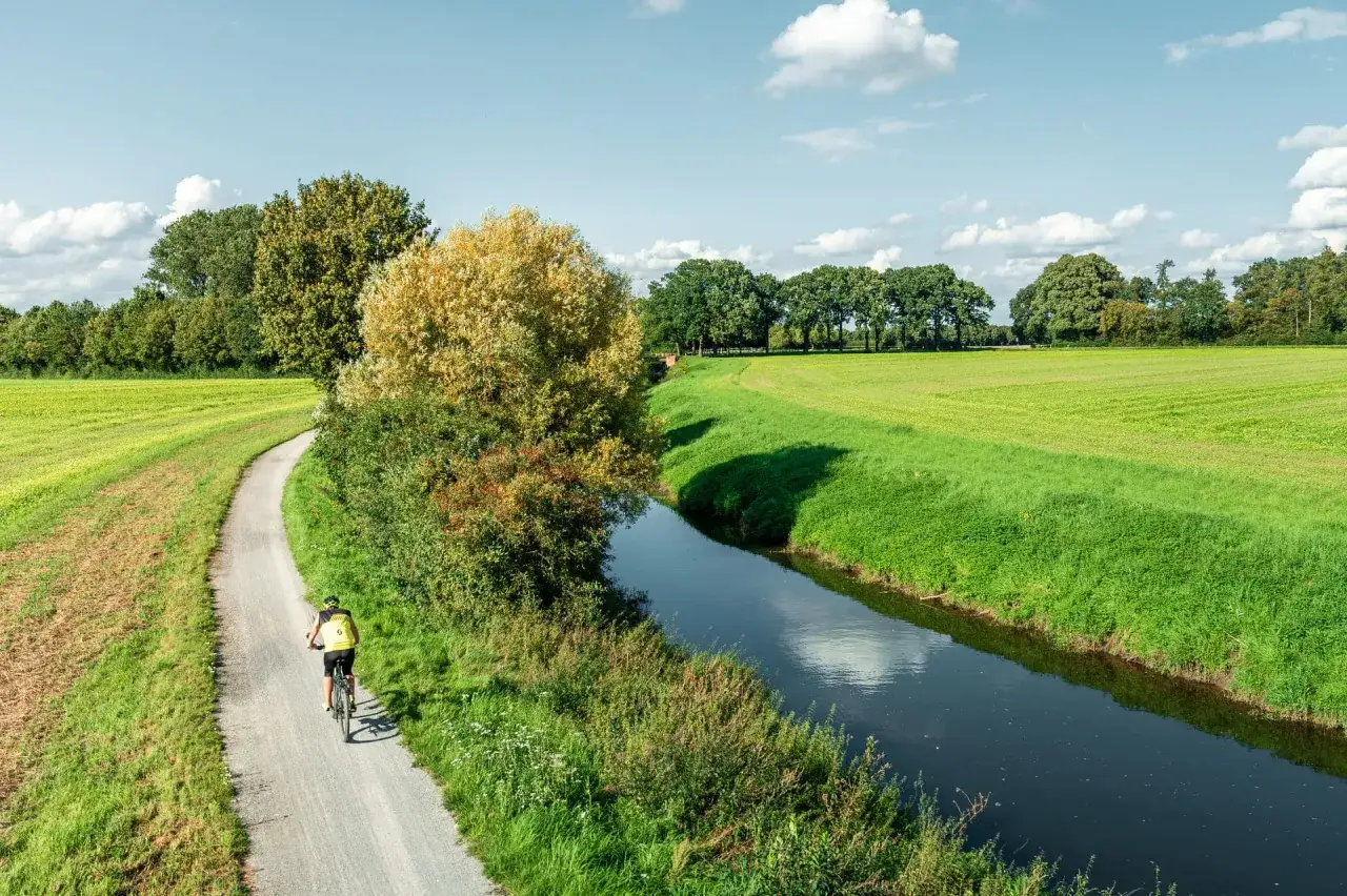 Radfahrer auf dem werse ems radweg, vorbei an grünen Feldern und einem Fluss unter blauem Himmel mit Wolken.