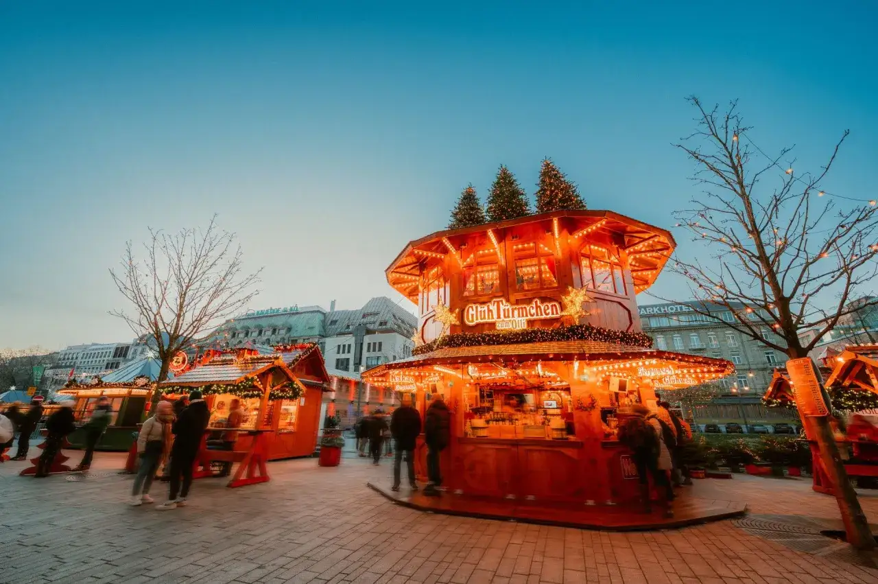 Glühweinbuden auf einem Weihnachtsmarkt in NRW, beleuchtet von Lichterketten. Menschen schlendern durch die festliche Atmosphäre.