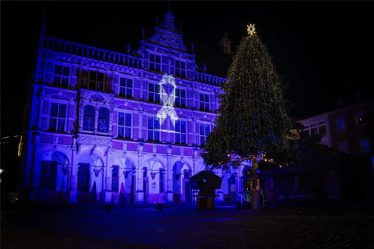 Festlicher Weihnachtsmarkt Bocholt: Ein prächtiger, beleuchteter Weihnachtsbaum steht vor dem Rathaus, das in blaues Licht getaucht ist und eine weiße Schleife projiziert.