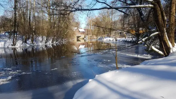 Winterliche Idylle an der **Planwiese Telgte**: Eisiger Fluss, schneebedeckte Ufer und ein Fachwerkhaus spiegeln sich im Wasser.