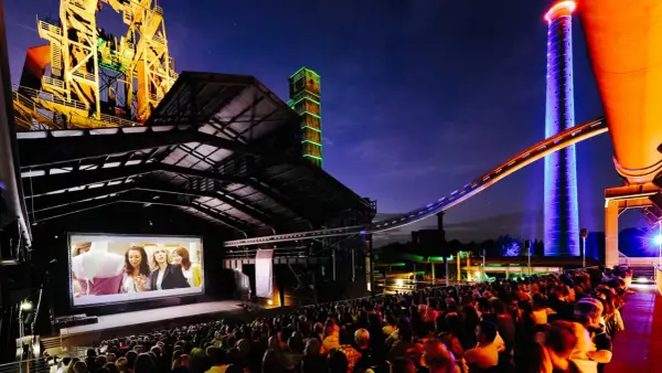 Ein belebtes **freiluft kino** mit vielen Zuschauern, die einen Film auf einer großen Leinwand vor einer beeindruckenden Industriekulisse mit bunten Türmen und Stahlkonstruktionen genießen.