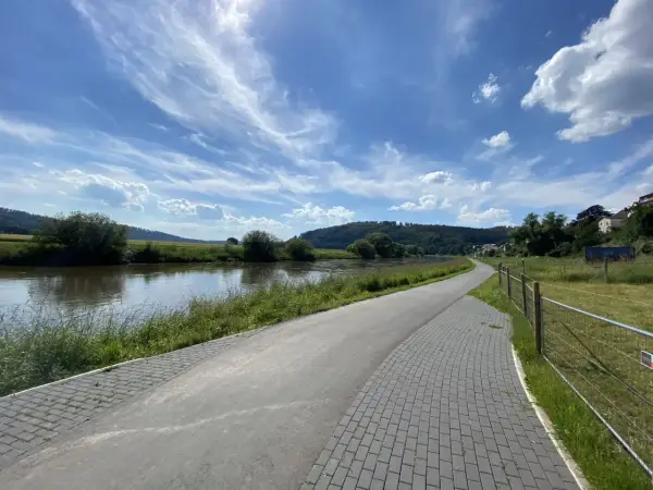 Ein asphaltierter Weg säumt den Fluss, ideal für eine Weser-Radweg Rundtour. Grüne Ufer und ein blauer Himmel mit Wolken laden zum Radfahren ein.