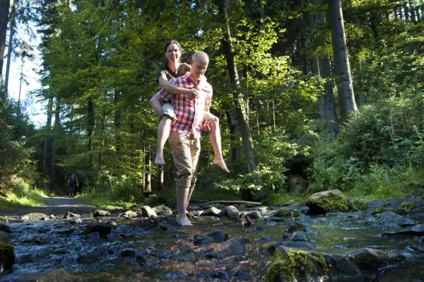 Ein Mann trägt eine Frau auf dem Rücken durch einen Bach im Wald. Die Sonne scheint durch die Bäume. Ein idyllischer Ort, wie eine teuto quelle.