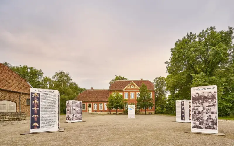 Das Haus Nottbeck mit Ausstellungen im Freien. Mehrere Schilder mit Fotos und Texten stehen vor dem roten Backsteingebäude.