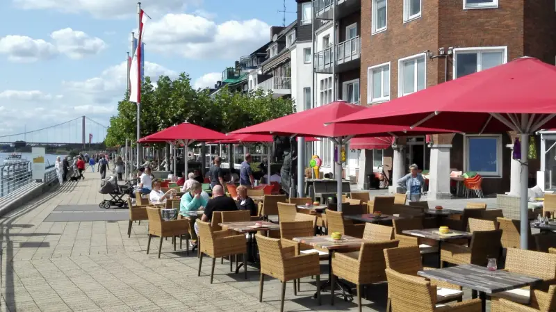 Gemütliche Straßencafés mit roten Sonnenschirmen laden zum Verweilen ein. Menschen spazieren am Rhein entlang, im Hintergrund die Brücke von Emmerich.
