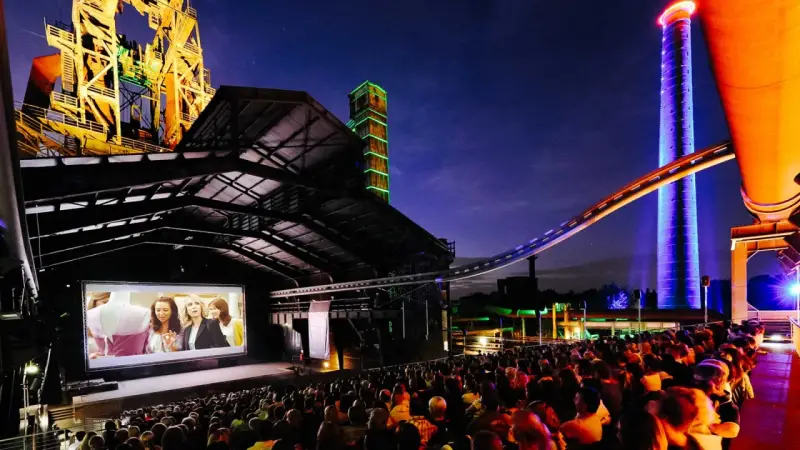 Ein belebtes **freiluft kino** mit vielen Zuschauern, die einen Film auf einer großen Leinwand vor einer beeindruckenden Industriekulisse mit bunten Türmen und Stahlkonstruktionen genießen.